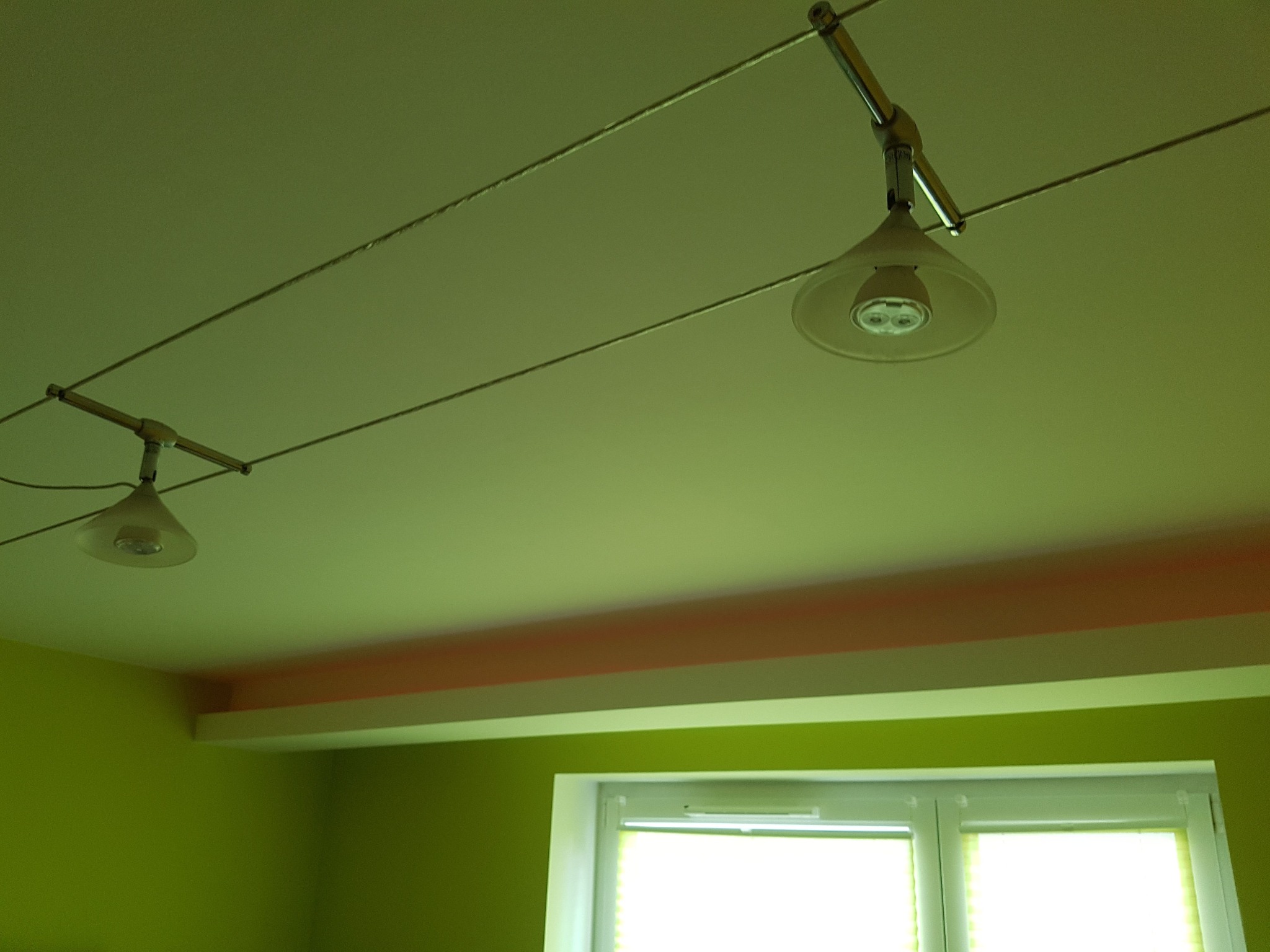 Dwa reflektory szynowe z przezroczystymi kloszami i żarówkami LED, zamontowane na suficie w pokoju z zielonymi ścianami i oknem.