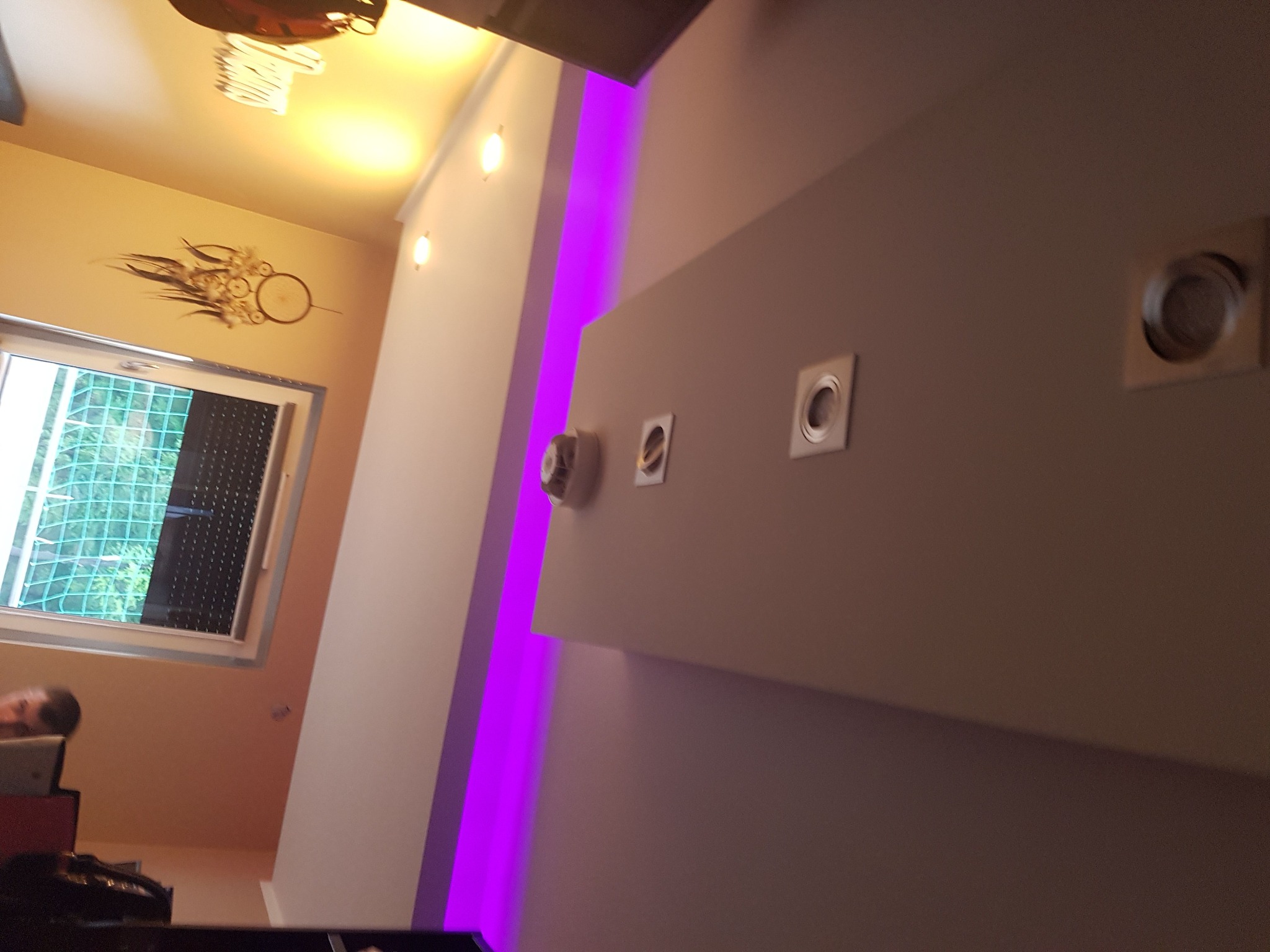 Nowoczesne wnętrze z podświetleniem LED w kolorze fioletowym, widoczne punktowe oświetlenie sufitowe i dekoracja na ścianie w odcieniach beżu.