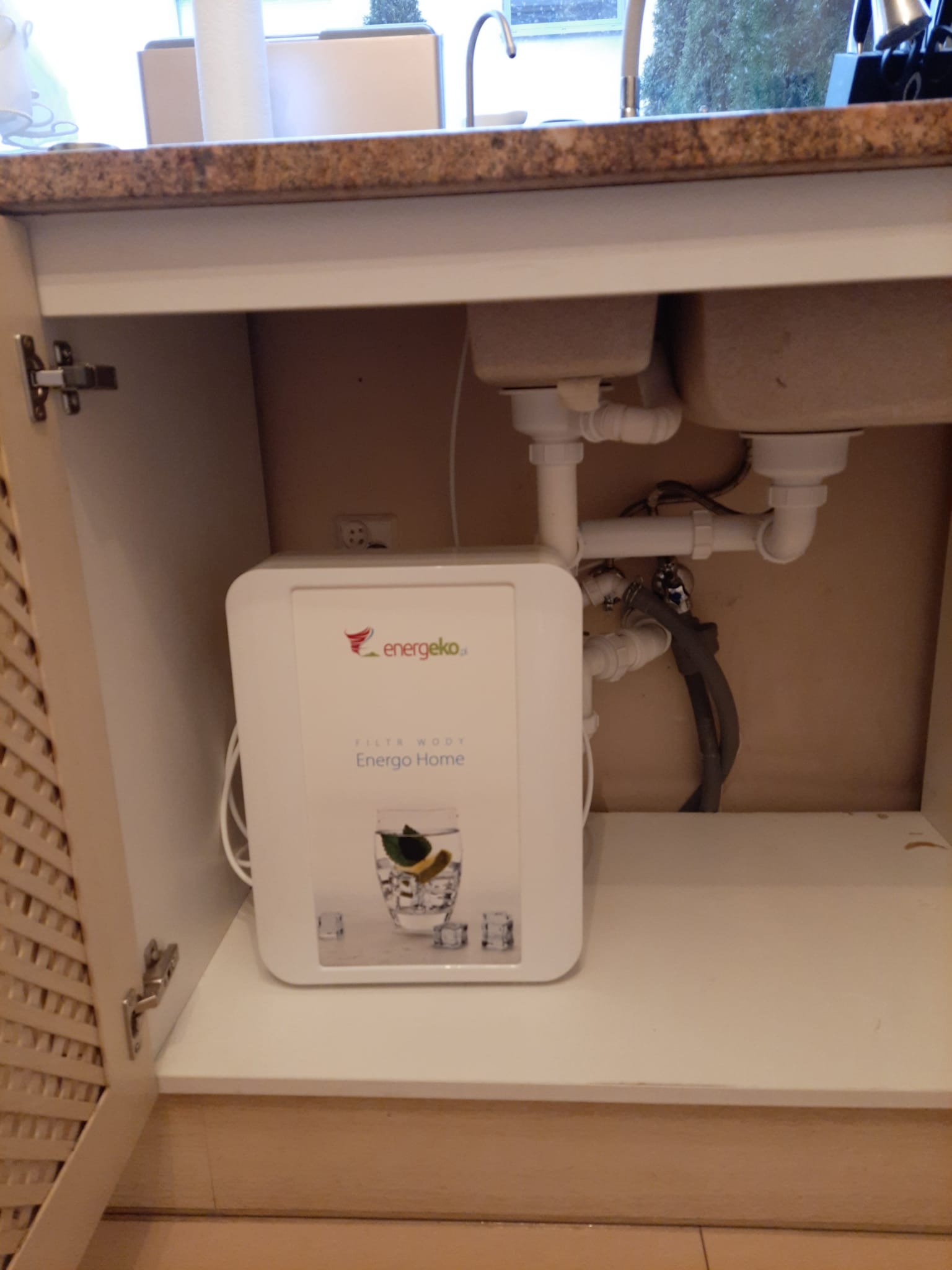 Kompaktowy filtr wody marki Energeko zamontowany w szafce pod zlewem, widoczne rury i syfon zlewozmywaka.
