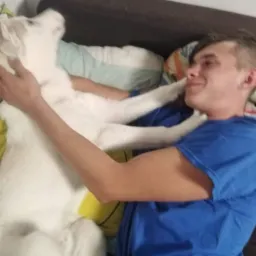 Młody mężczyzna w niebieskiej koszulce leży na kanapie, obejmując dużego, białego psa rasy husky.