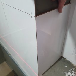 Kistela Usługi Budowlane - Proces układania białych, błyszczących płytek ściennych z użyciem poziomicy laserowej i krzyżyków dystansowych, widoczny fragment dłoni glazurnika.