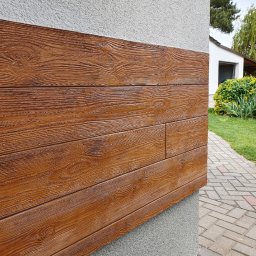 Kistela Usługi Budowlane - Elewacja budynku z dekoracyjnymi panelami imitującymi drewno, z widoczną fakturą słojów, na tle szarego tynku strukturalnego i chodnika z kostki brukowej.