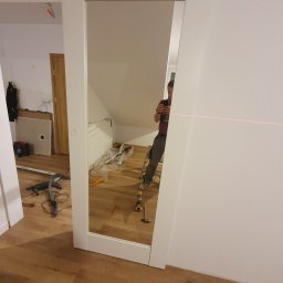 Kistela Usługi Budowlane - Montaż przesuwnych drzwi z lustrem w poddaszowym pokoju, widoczny laserowy wskaźnik poziomu, narzędzia i odblask osoby robiącej zdjęcie.