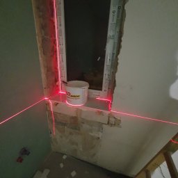Kistela Usługi Budowlane - Przygotowanie do osadzania okna z użyciem poziomicy laserowej; widoczny surowy mur, wiadro z zaprawą i fragment podłogi z resztkami materiałów budowlanych.