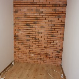 Kistela Usługi Budowlane - Ściana wyłożona cegłą klinkierową w małym pomieszczeniu, widoczne przewody elektryczne i sprzęt budowlany na podłodze, surowe wykończenie.