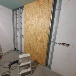 Kistela Usługi Budowlane - Montaż płyty OSB na metalowym stelażu, widoczny laserowy wskaźnik poziomu, drabinka, wiertarka udarowa i częściowo zamontowana płyta gipsowo-kartonowa na suficie.