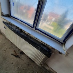 Kistela Usługi Budowlane - Okno zabezpieczone folią i taśmą malarską podczas prac remontowych, grzejnik osłonięty czarną folią, widoczne resztki pianki montażowej na parapecie i podłodze.