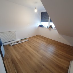 Kistela Usługi Budowlane - Poddasze z ułożonymi panelami podłogowymi w odcieniach brązu, widoczne opakowania paneli przy ścianie, grzejnik, okno z roletami, surowe białe ściany i żarówka na suficie.