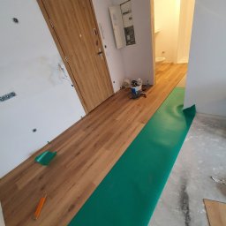 Kistela Usługi Budowlane - W trakcie układania paneli podłogowych w jasnym wnętrzu; widoczny podkład w kolorze zielonym, docięte panele, narzędzia oraz fragment łazienki w tle.