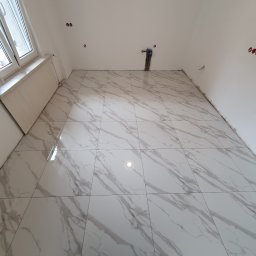 Kistela Usługi Budowlane - Ściana łazienki wyłożona błyszczącymi, białymi płytkami imitującymi marmur, widoczne przygotowane punkty instalacji wodno-kanalizacyjnej i elektrycznej, surowy stan wykończenia.