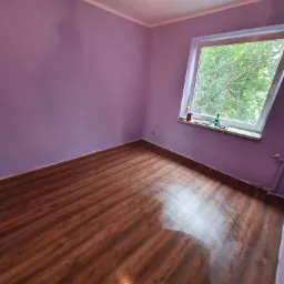 Pokój po remoncie: świeżo pomalowane liliowe ściany, panele podłogowe z efektem drewna, okno dachowe z widokiem na zieleń.