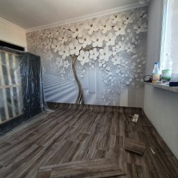 Kistela Usługi Budowlane - Pokój w trakcie remontu z nową podłogą imitującą drewno, fragmenty paneli leżące na podłodze, ściana z fototapetą przedstawiającą drzewo z białymi kwiatami, zabezpieczone meble folią malarską.