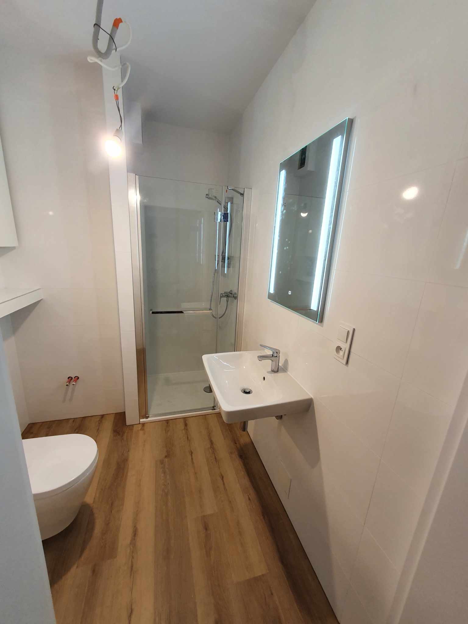 Nowoczesna łazienka z kabiną prysznicową, umywalką nablatową z podświetlanym lustrem, wykończona białymi płytkami i panelami podłogowymi w odcieniach drewna; widoczne niedokończone podłączenie...