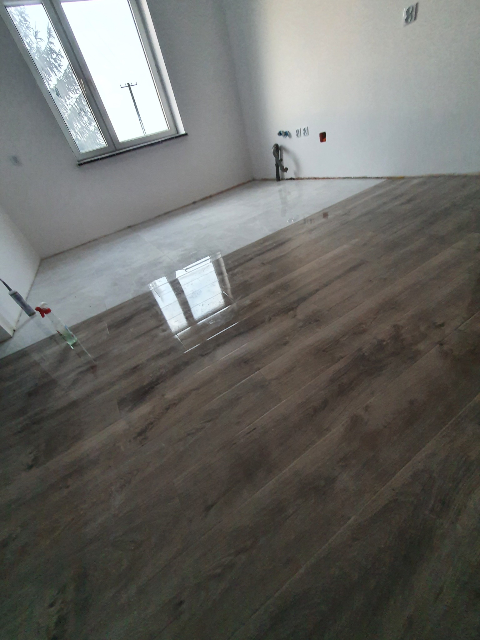 Pokój w trakcie prac wykończeniowych: ułożone panele podłogowe z ciemnego drewna kontrastują z fragmentem podłogi przygotowanej pod wylewkę, widoczne okno dachowe i instalacje na ścianie.