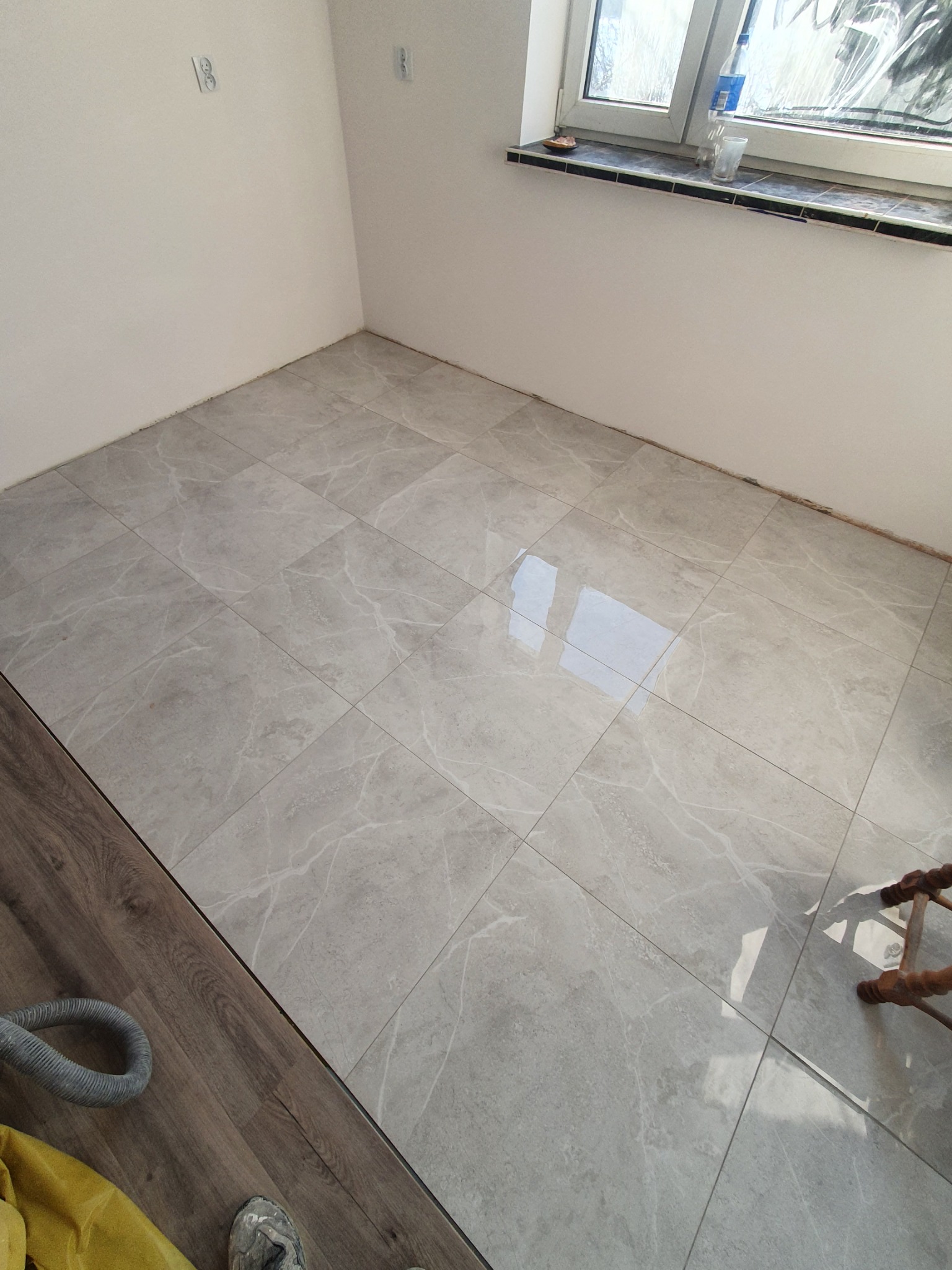 Świeżo ułożona, jasnoszara podłoga z płytek imitujących marmur, kontrastująca z fragmentem ciemnobrązowych paneli podłogowych i wężem od odkurzacza w rogu pomieszczenia, widok na okno z parapetem...