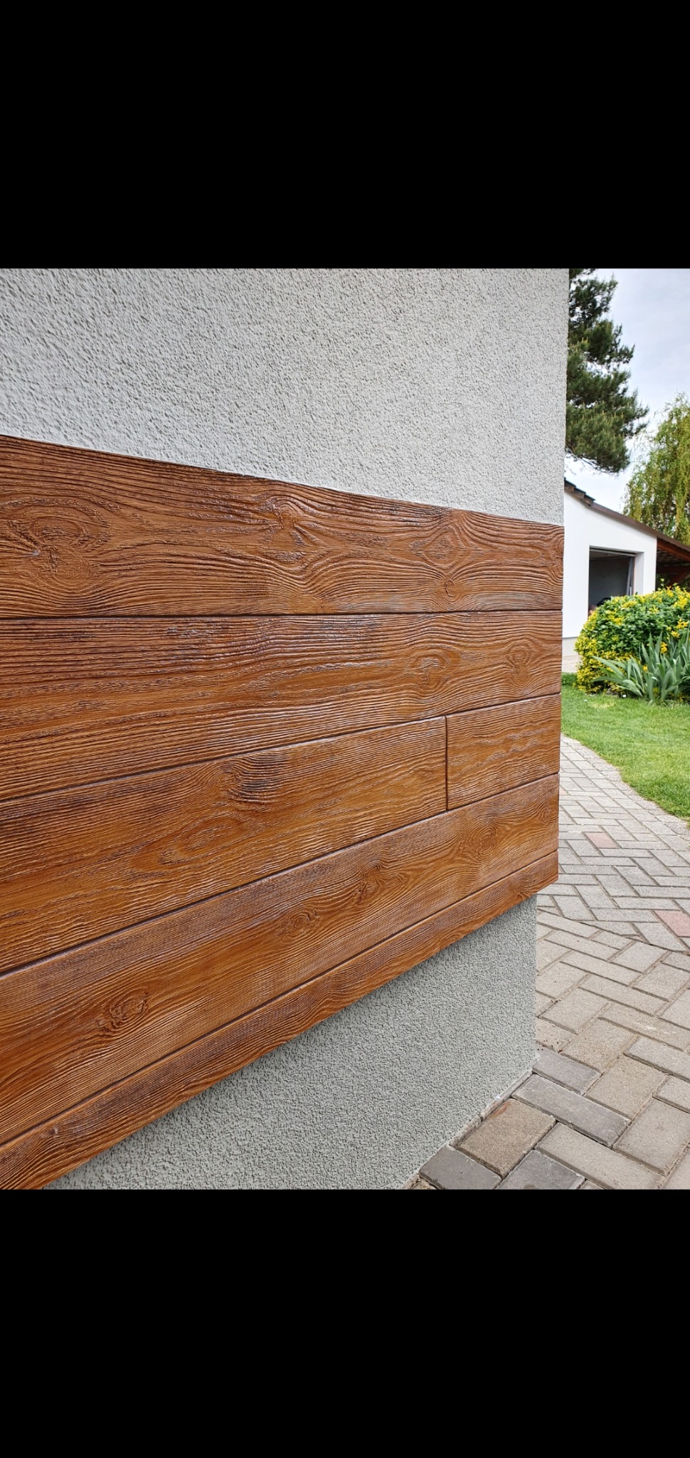 Elewacja budynku z dekoracyjnymi panelami imitującymi drewno, z widoczną fakturą słojów, na tle szarego tynku strukturalnego i chodnika z kostki brukowej.