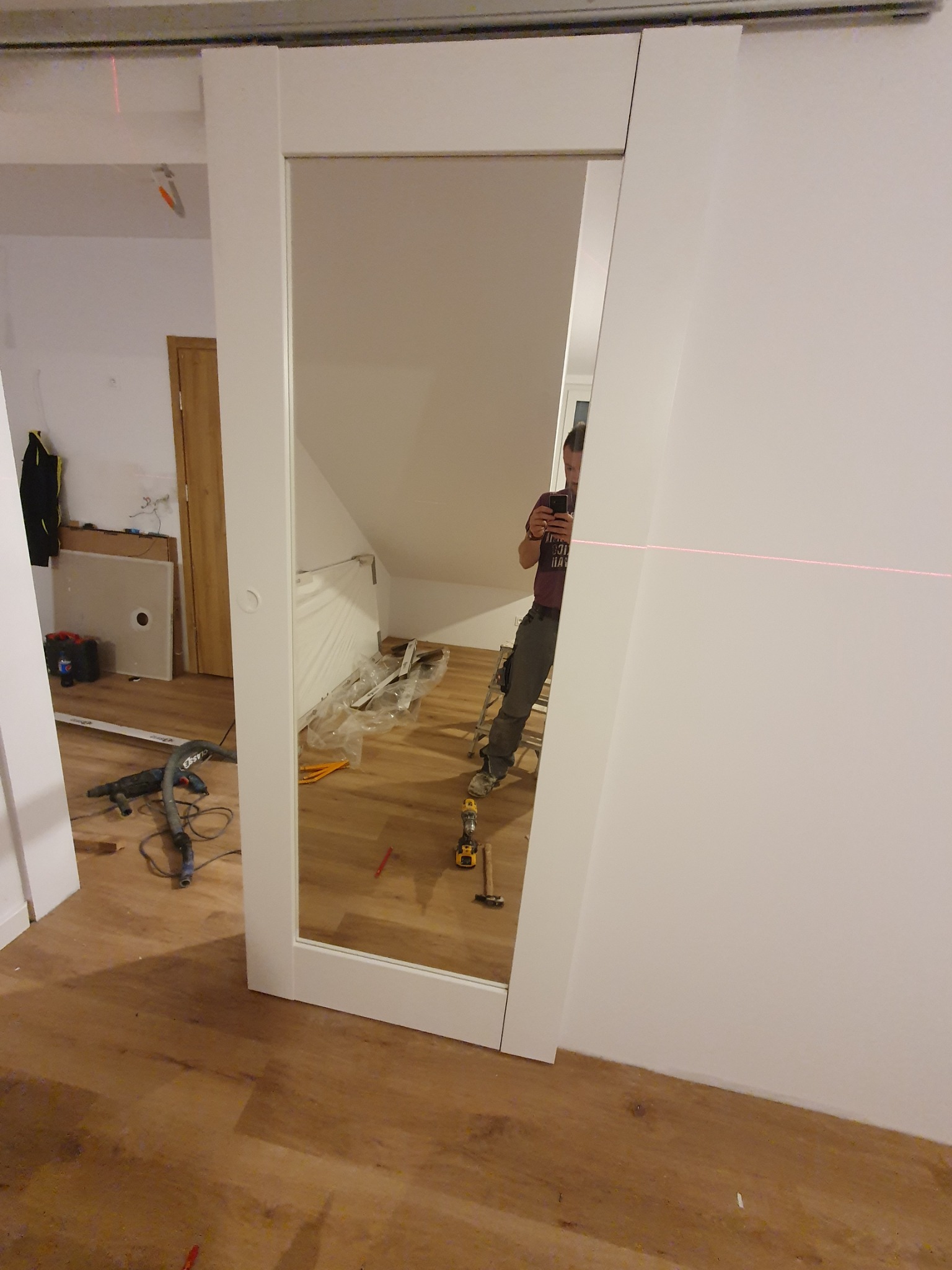 Montaż przesuwnych drzwi z lustrem w poddaszowym pokoju, widoczny laserowy wskaźnik poziomu, narzędzia i odblask osoby robiącej zdjęcie.