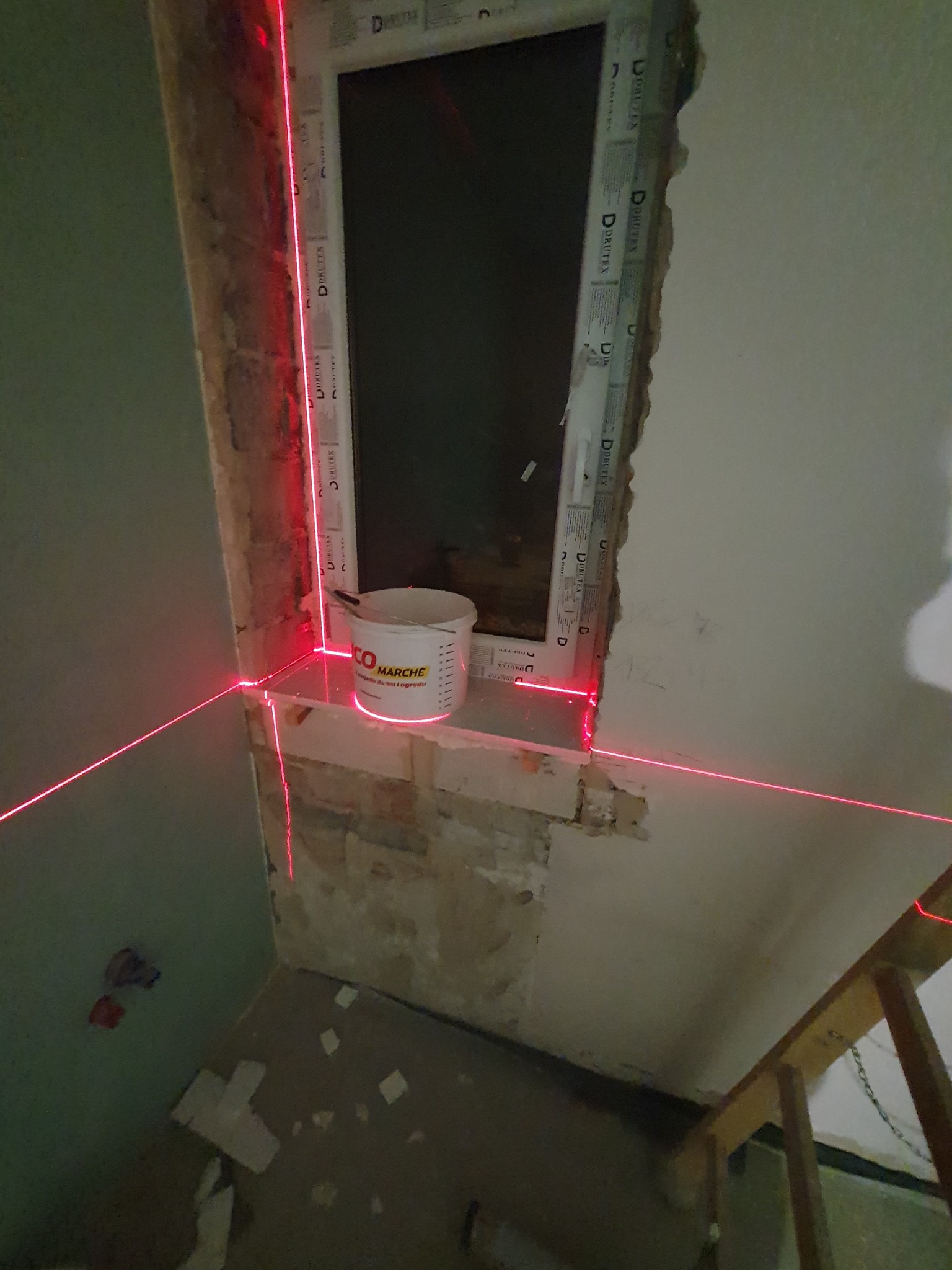 Przygotowanie do osadzania okna z użyciem poziomicy laserowej; widoczny surowy mur, wiadro z zaprawą i fragment podłogi z resztkami materiałów budowlanych.
