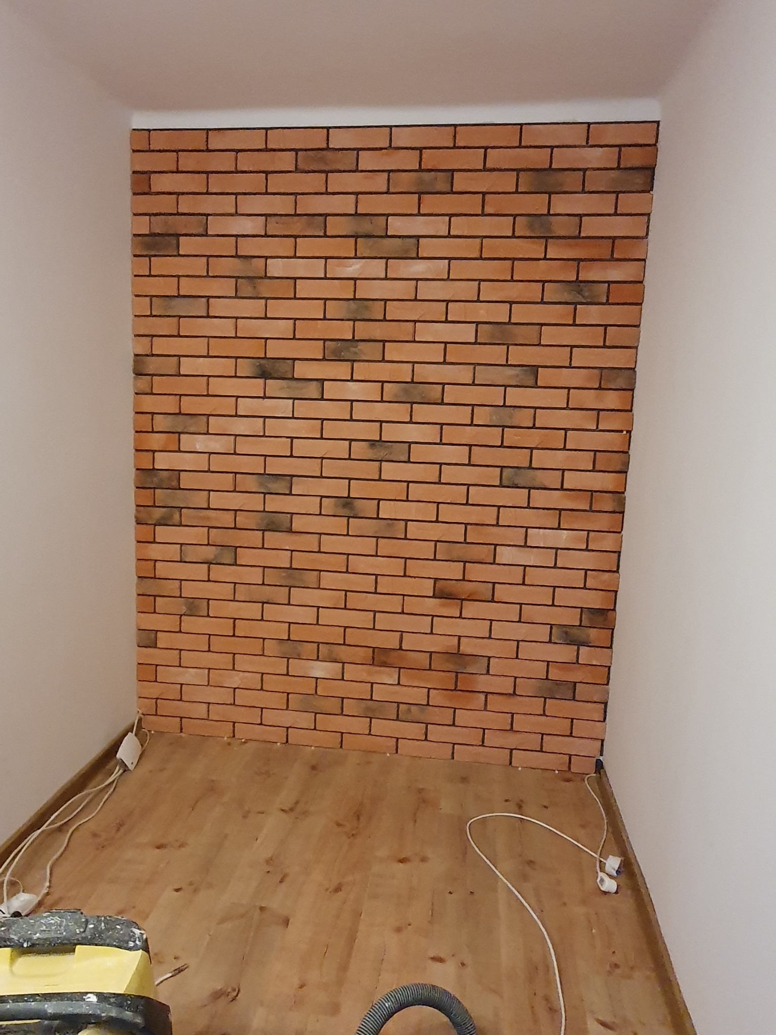 Ściana wyłożona cegłą klinkierową w małym pomieszczeniu, widoczne przewody elektryczne i sprzęt budowlany na podłodze, surowe wykończenie.