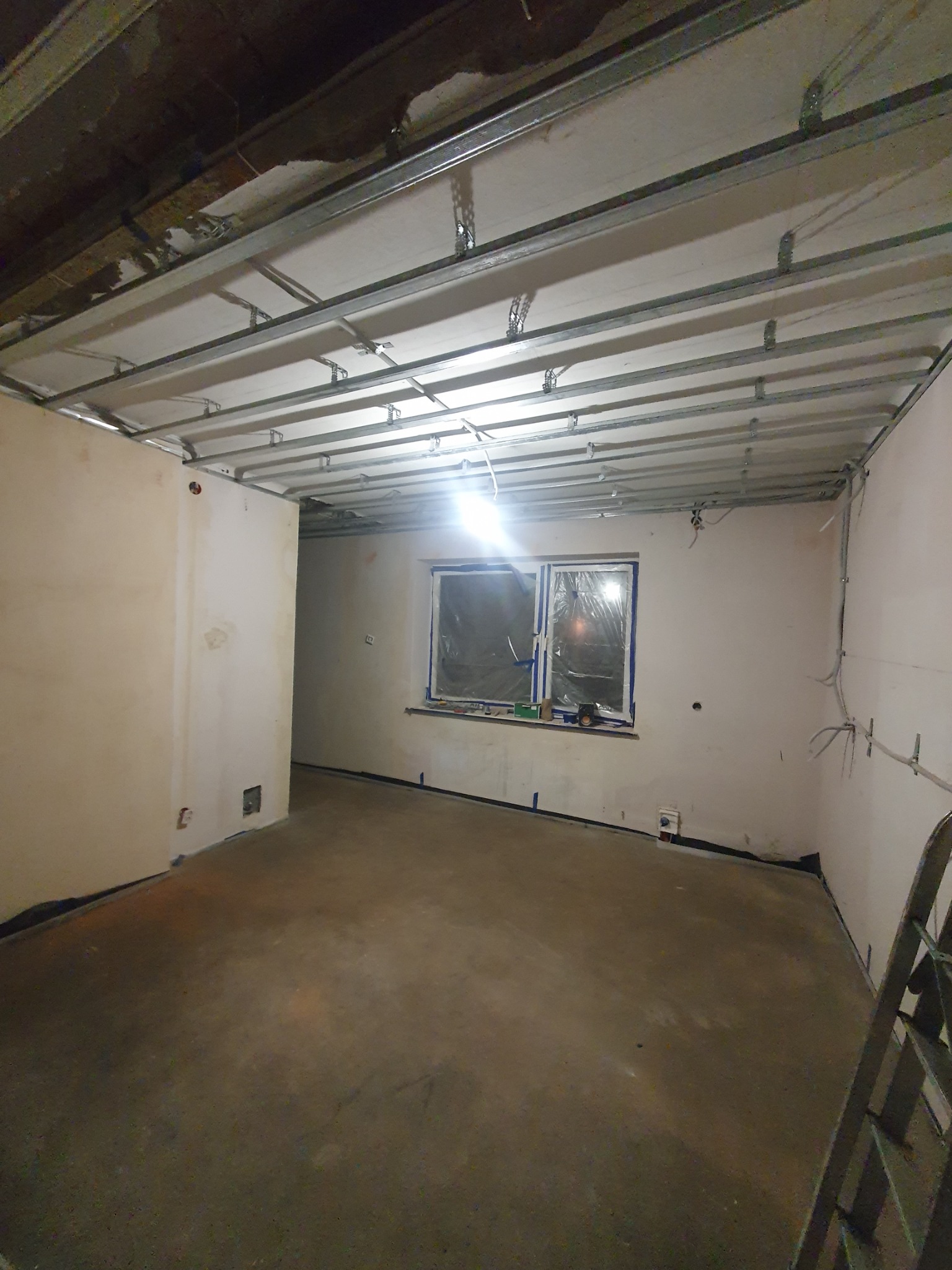 Wnętrze w trakcie prac remontowych: widoczne metalowe profile sufitowe, surowe ściany z widocznymi przewodami elektrycznymi, okno zabezpieczone folią malarską, surowa podłoga, drabina w rogu...