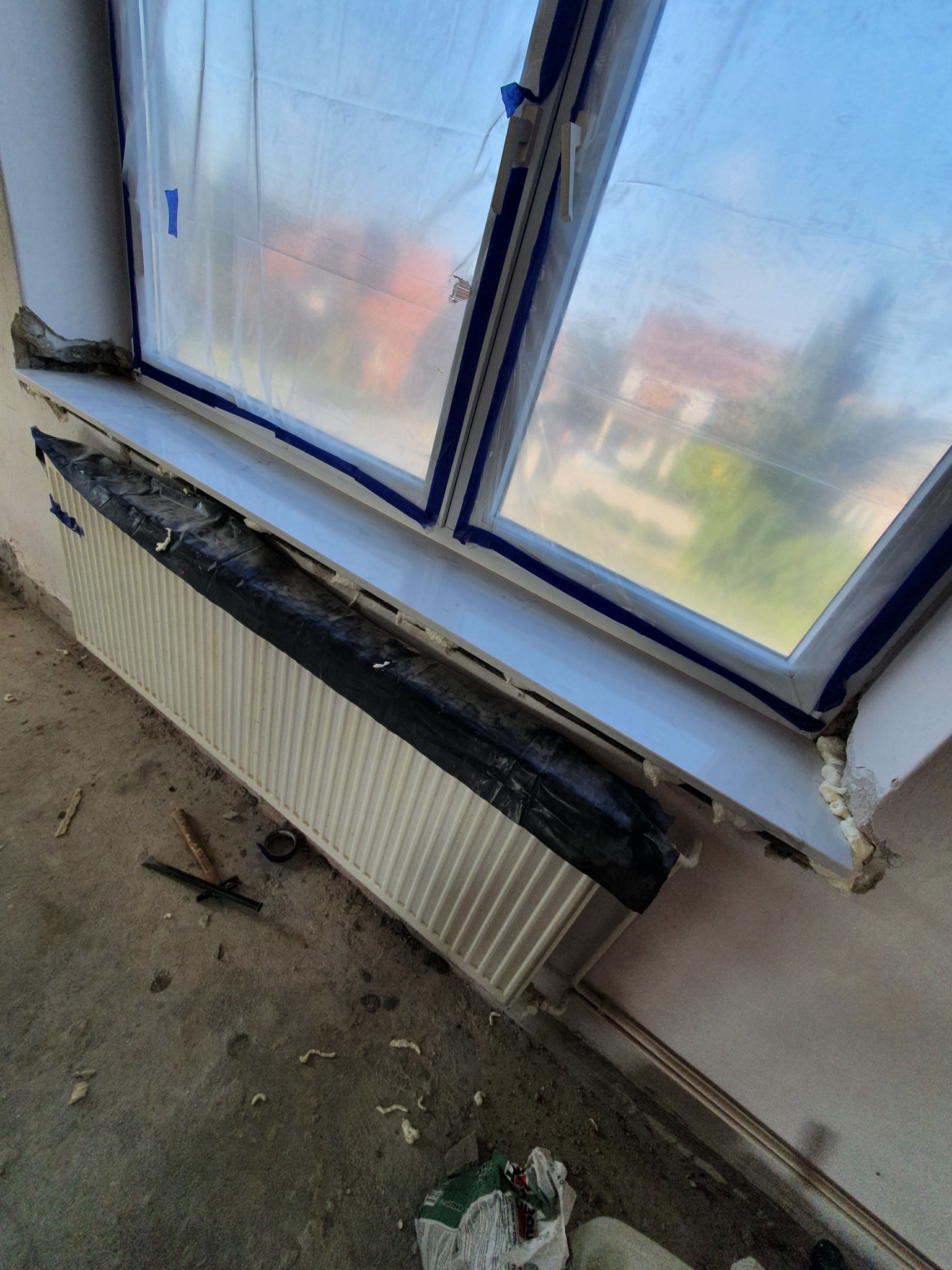 Okno zabezpieczone folią i taśmą malarską podczas prac remontowych, grzejnik osłonięty czarną folią, widoczne resztki pianki montażowej na parapecie i podłodze.