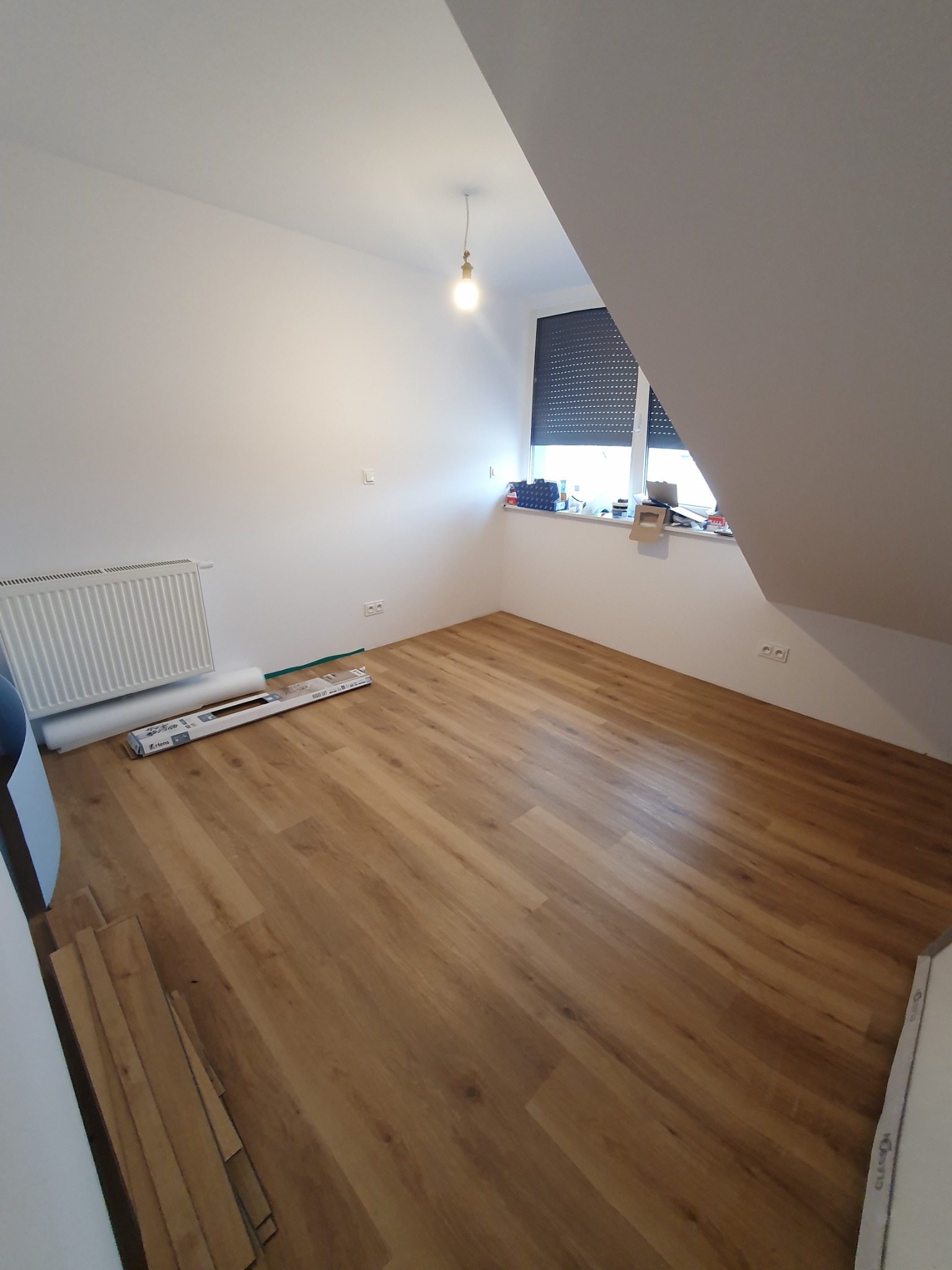 Poddasze z ułożonymi panelami podłogowymi w odcieniach brązu, widoczne opakowania paneli przy ścianie, grzejnik, okno z roletami, surowe białe ściany i żarówka na suficie.