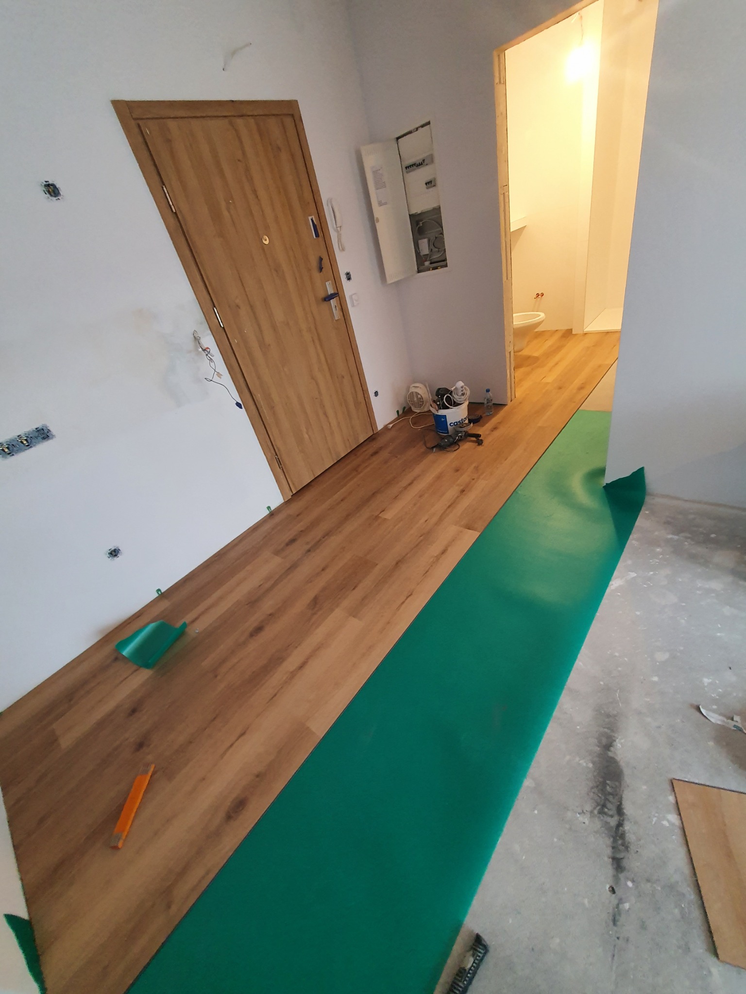 W trakcie układania paneli podłogowych w jasnym wnętrzu; widoczny podkład w kolorze zielonym, docięte panele, narzędzia oraz fragment łazienki w tle.