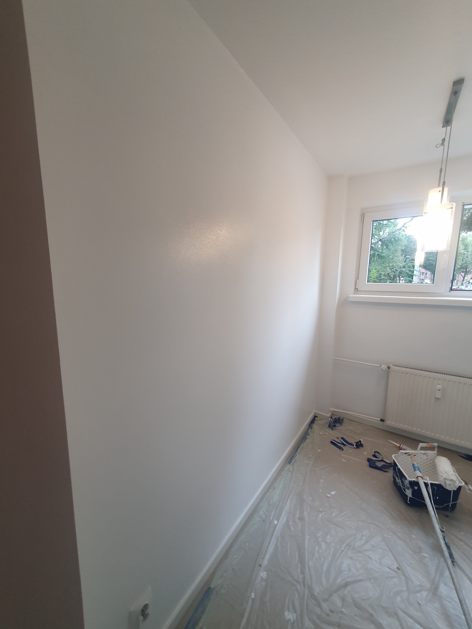 Świeżo pomalowany pokój na biało, widoczne narzędzia malarskie, folia ochronna na podłodze, okno z widokiem na zieleń.