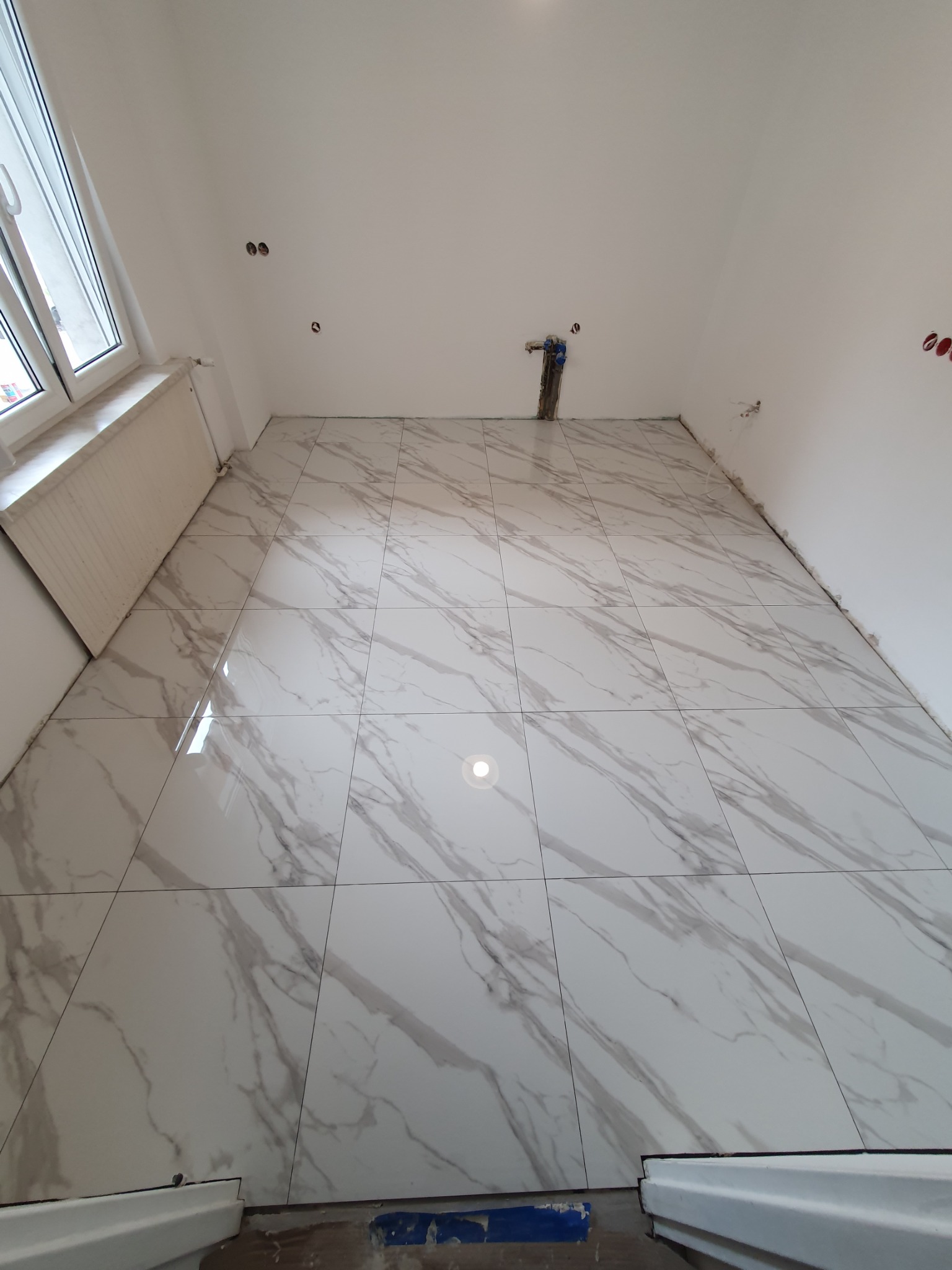 Ściana łazienki wyłożona błyszczącymi, białymi płytkami imitującymi marmur, widoczne przygotowane punkty instalacji wodno-kanalizacyjnej i elektrycznej, surowy stan wykończenia.