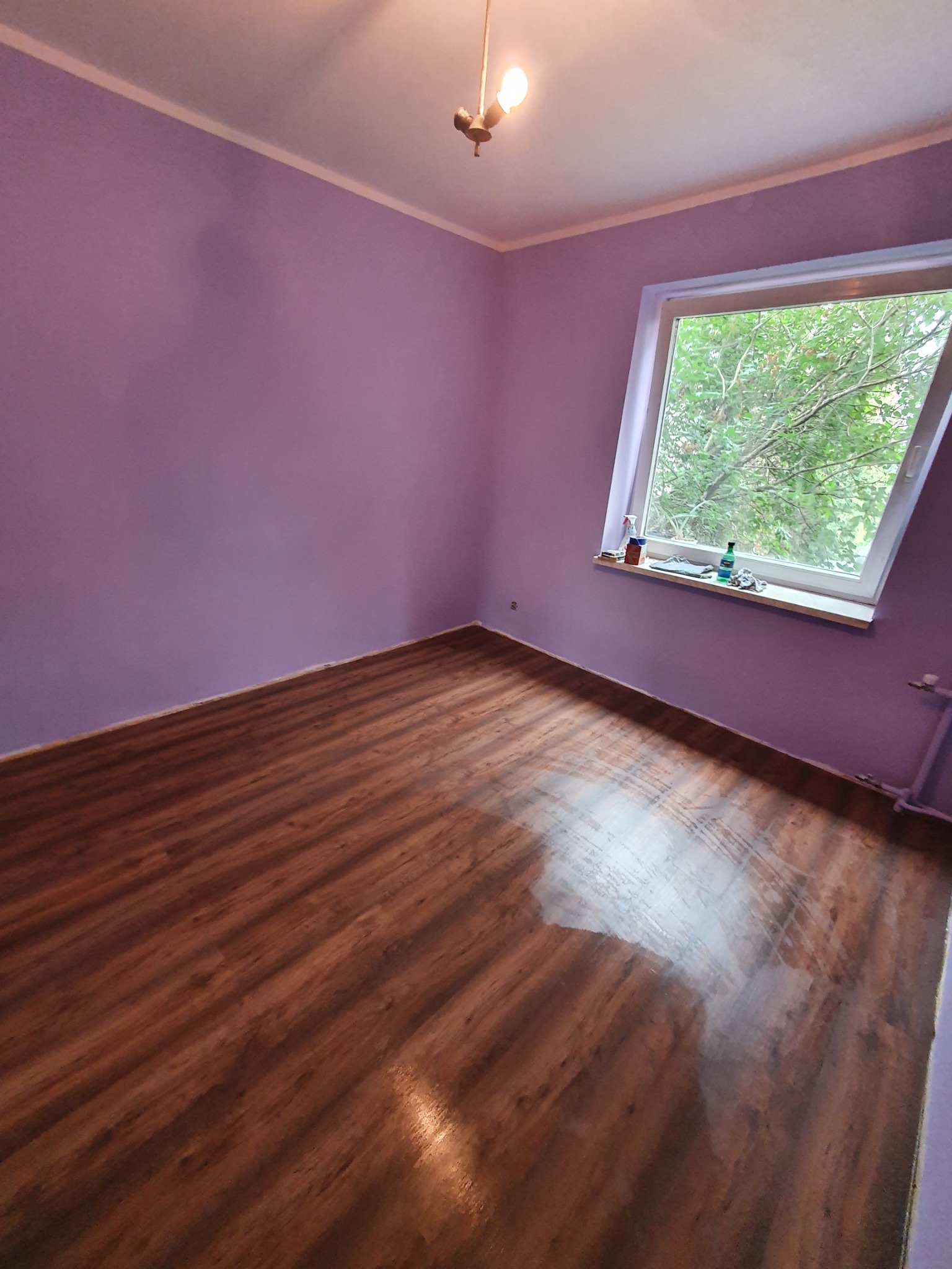 Pokój po remoncie: świeżo pomalowane liliowe ściany, panele podłogowe z efektem drewna, okno dachowe z widokiem na zieleń.