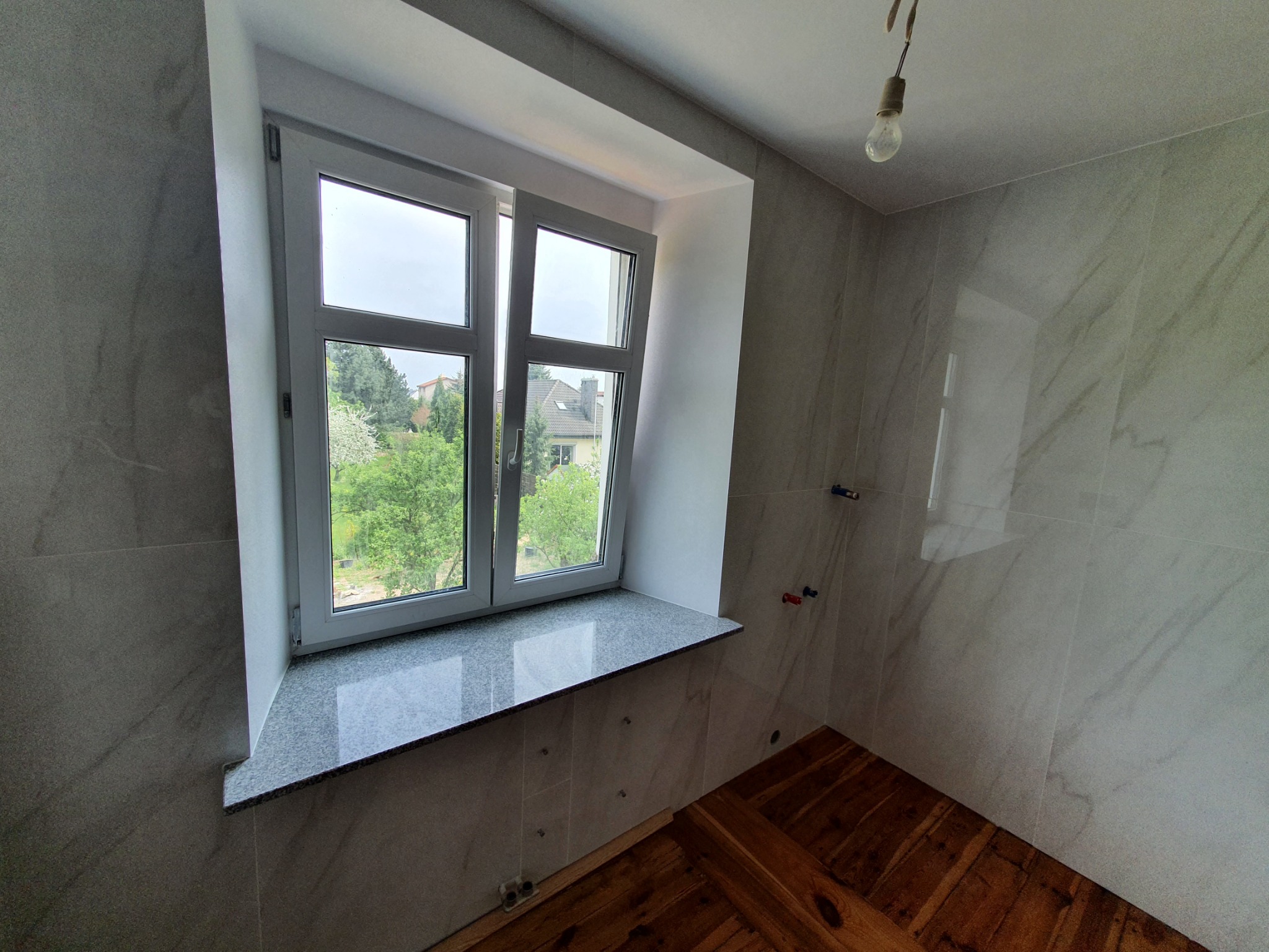 Wykończona łazienka z marmurowymi płytkami ściennymi, drewnianą podłogą i oknem z kamiennym parapetem, widok na zieleń za oknem, surowa żarówka zwisająca z sufitu.