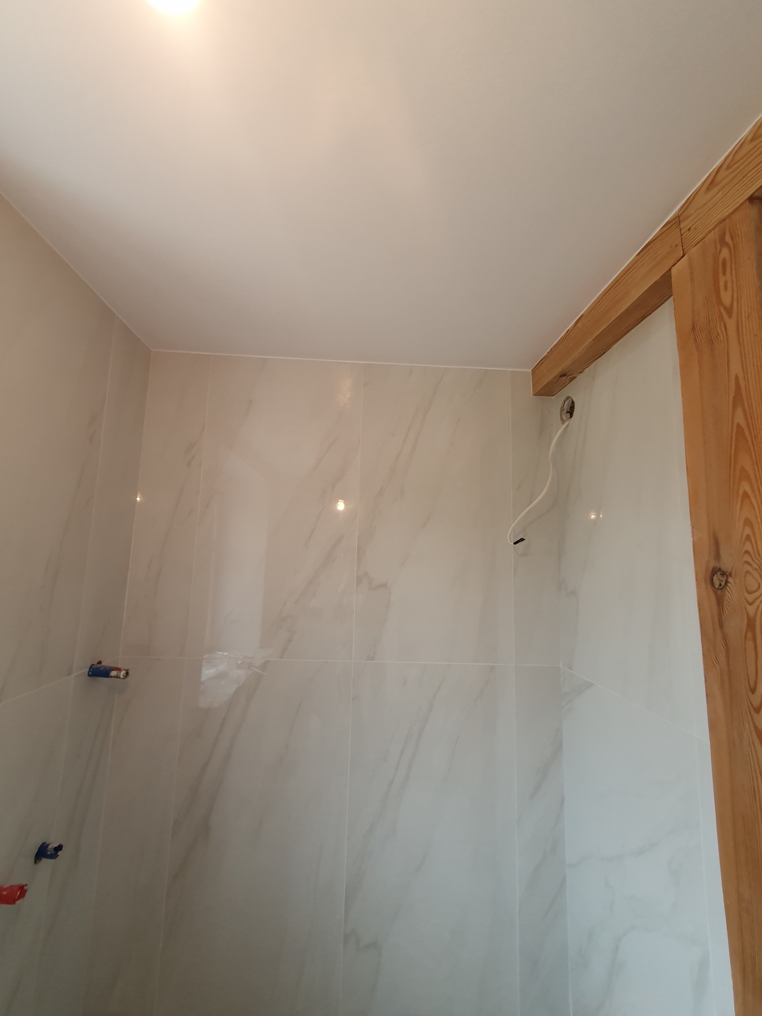 Wnętrze łazienki w trakcie remontu, widoczne płytki imitujące marmur na ścianach, drewniana rama drzwi, wystające przewody elektryczne i rury instalacji wodnej.