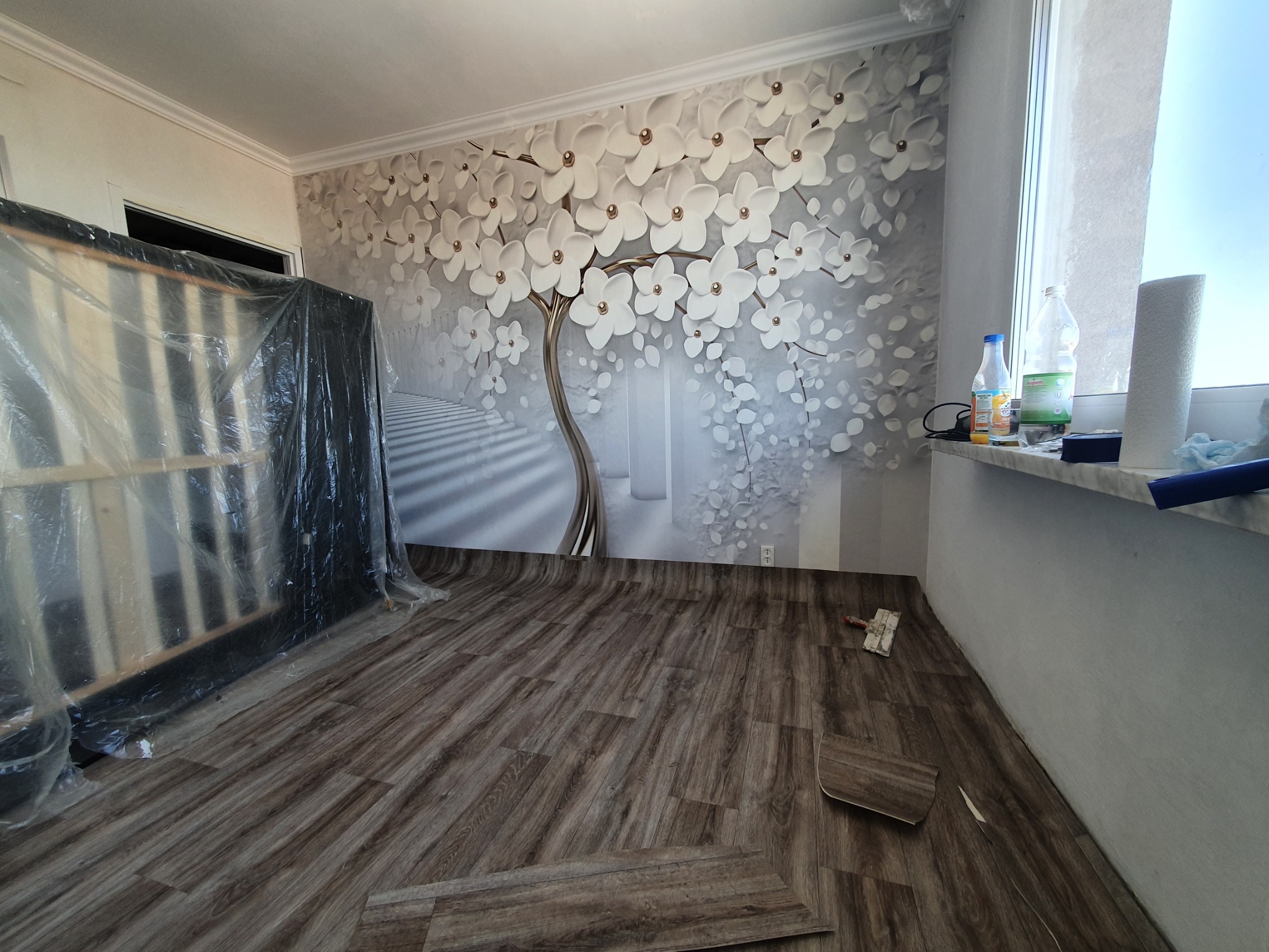 Pokój w trakcie remontu z nową podłogą imitującą drewno, fragmenty paneli leżące na podłodze, ściana z fototapetą przedstawiającą drzewo z białymi kwiatami, zabezpieczone meble folią malarską.