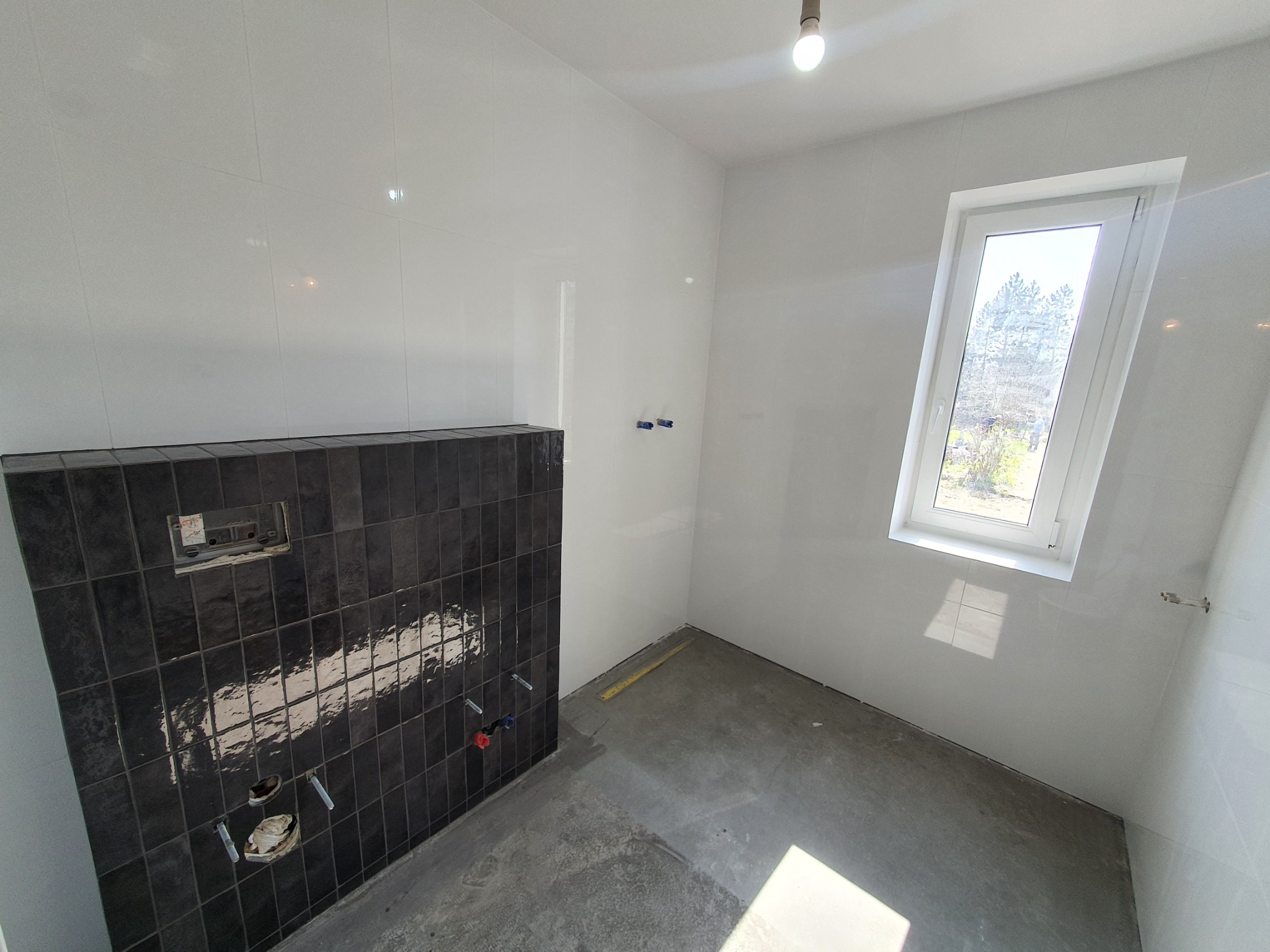 Wnętrze łazienki w trakcie remontu: ściany wyłożone białymi płytkami, fragment ściany z czarnymi płytkami, przygotowane instalacje wodno-kanalizacyjne, surowa betonowa podłoga, okno z widokiem...