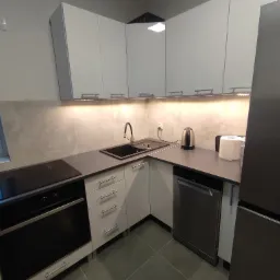 Nowoczesna kuchnia z białymi szafkami, szarym blatem i sprzętem AGD w zabudowie, w tym piekarnikiem, płytą indukcyjną i zmywarką, oświetlona listwą LED pod szafkami górnymi.