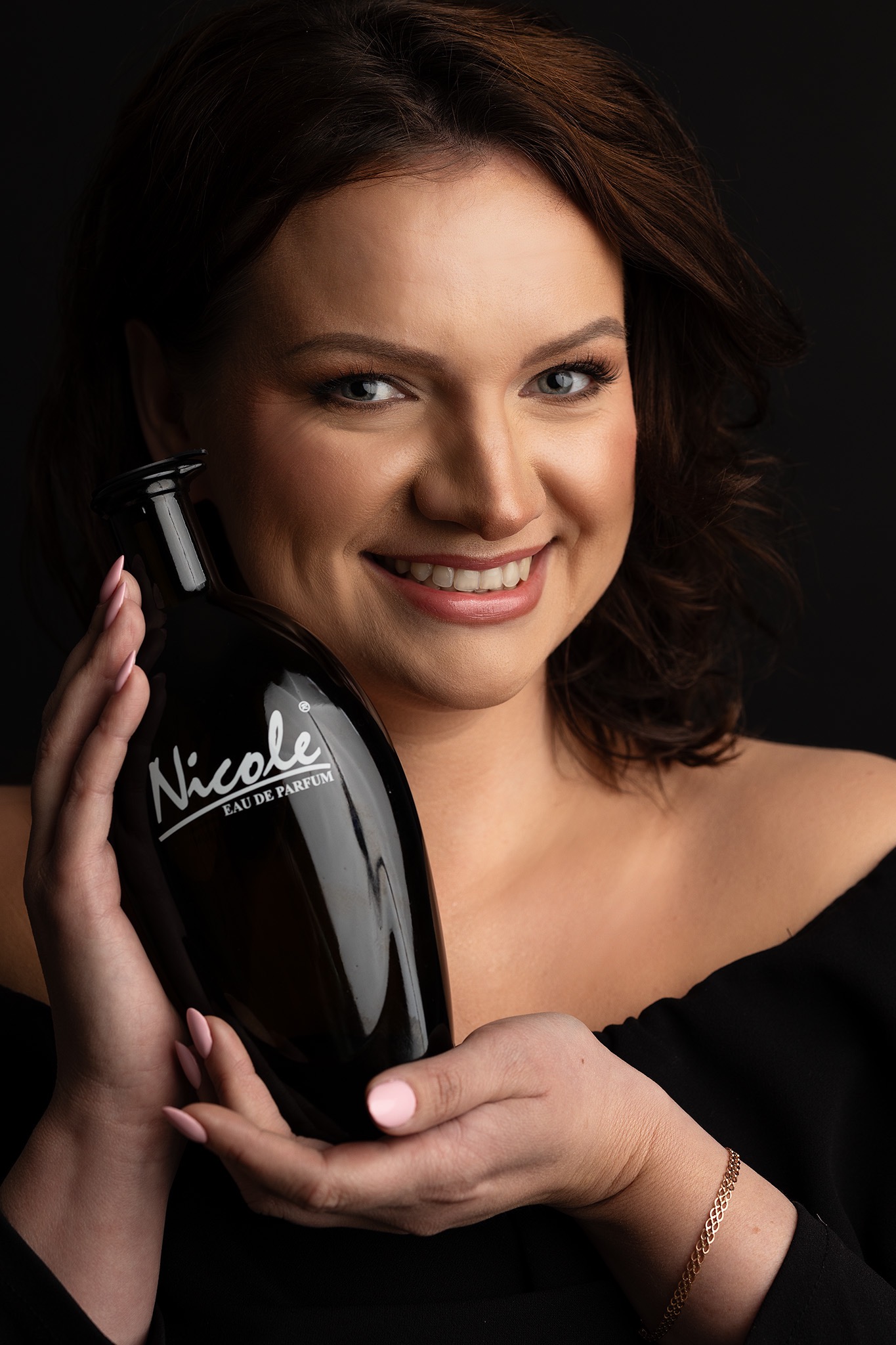 Uśmiechnięta kobieta prezentuje flakon perfum Nicole na czarnym tle, z delikatnym makijażem podkreślającym jej rysy twarzy i subtelnym różowym manicurem.