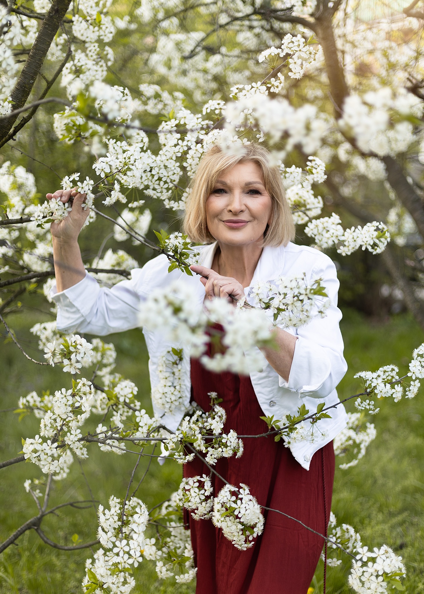 Uśmiechnięta blond kobieta w białej kurtce i czerwonej sukience, pozuje wśród kwitnących gałęzi drzewa owocowego w słoneczny dzień, z delikatnym makijażem podkreślającym rysy twarzy.