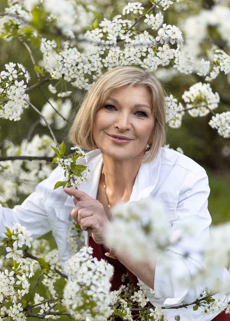 Uśmiechnięta kobieta z delikatnym makijażem, otoczona kwitnącymi gałązkami drzew owocowych, ubrana w białą koszulę.