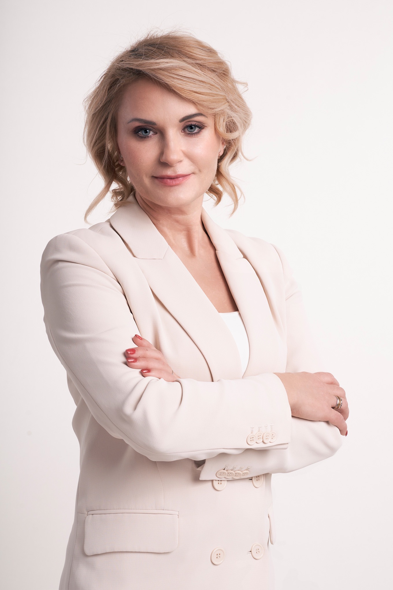 Portret kobiety o blond włosach i niebieskich oczach, w kremowym garniturze, z założonymi rękami, na jasnym tle.