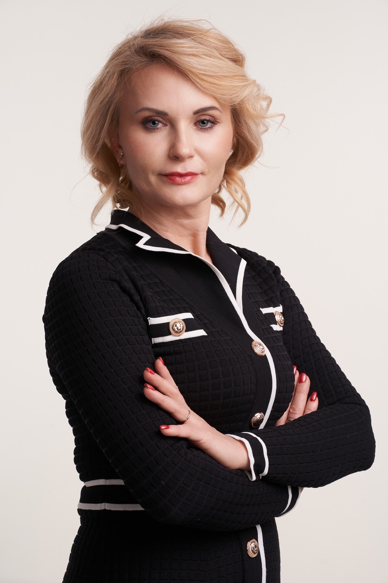 Portret kobiety z blond włosami, subtelnym makijażem i czerwonymi ustami, ubrana w elegancką czarną sukienkę z białymi lamówkami i złotymi guzikami.