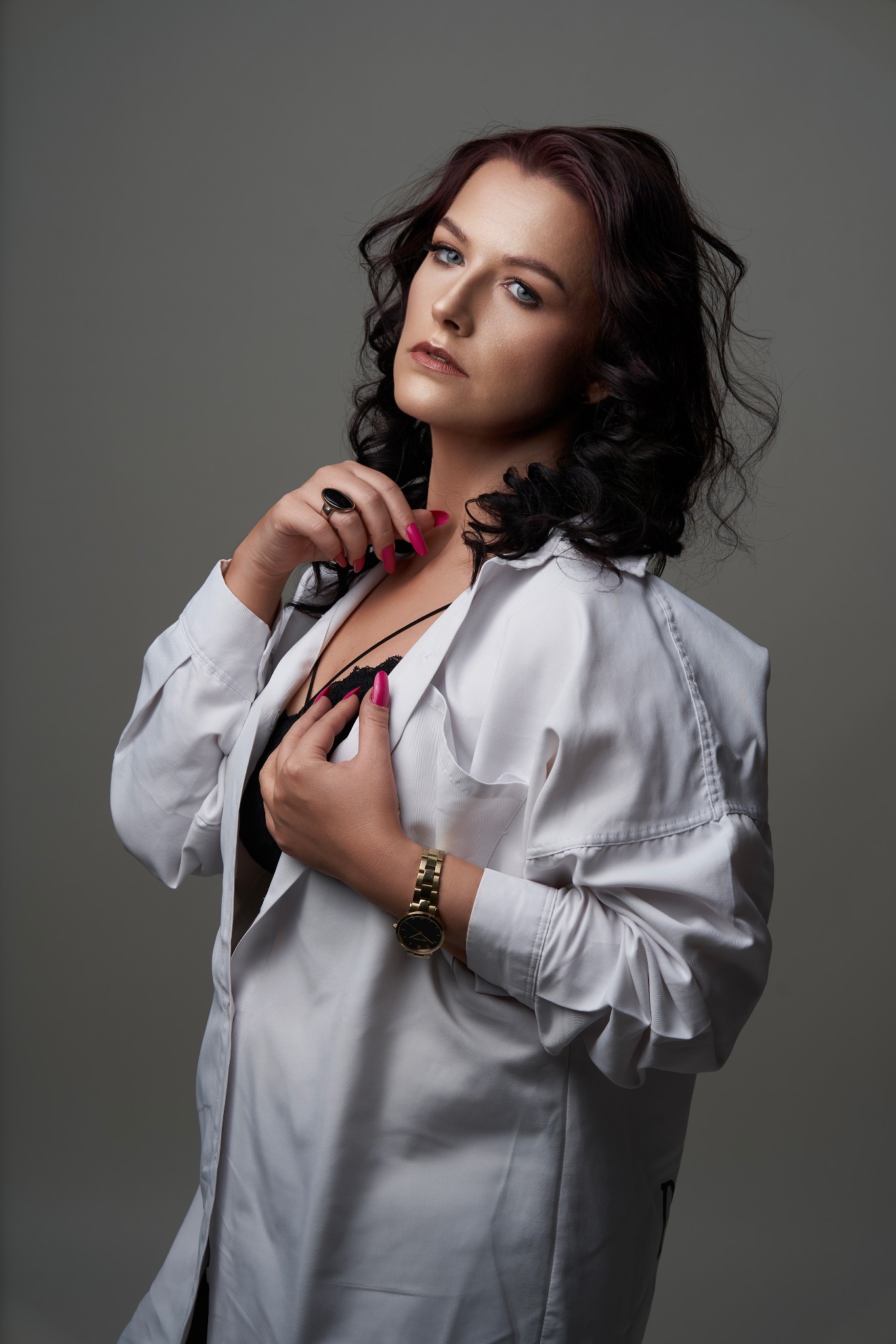 Portret kobiety z profesjonalnym, subtelnym makijażem, w rozpiętej białej koszuli, eksponującej czarną koronkową bieliznę, z ciemnymi, kręconymi włosami i złotym zegarkiem na ręce.