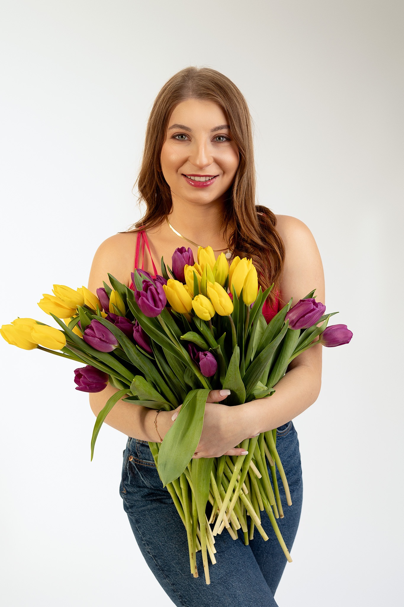 Uśmiechnięta kobieta z długimi brązowymi włosami, w dżinsach i różowej bluzce, trzyma bukiet żółtych i fioletowych tulipanów na białym tle.