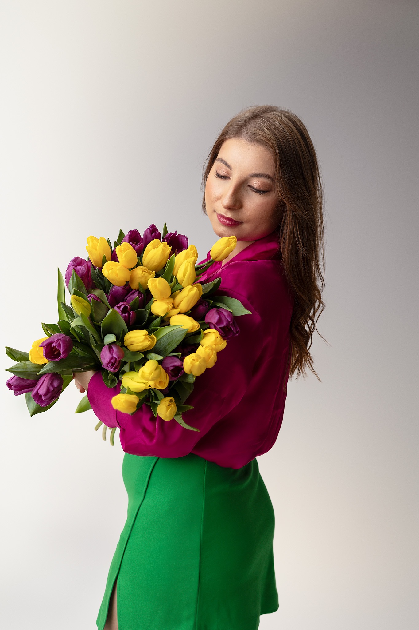 Kobieta z delikatnym makijażem ust, trzyma bukiet żółtych i fioletowych tulipanów, ubrana w fuksjową bluzkę i zieloną spódnicę.