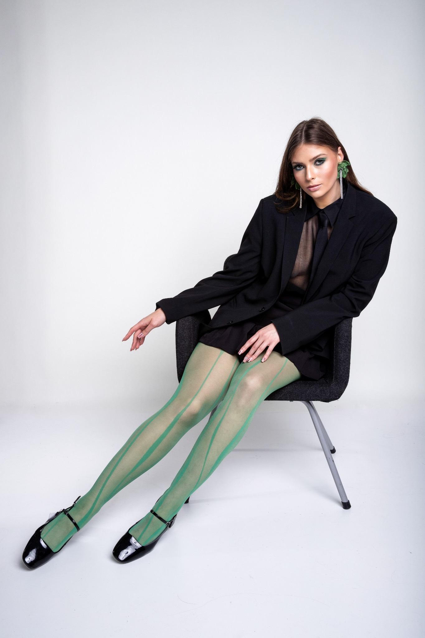 Portret kobiety w czarnym oversize'owym garniturze i zielonych rajstopach w paski, siedzącej na krześle na białym tle, z wyrazistym makijażem oczu i dużymi, wiszącymi kolczykami z zielonymi...