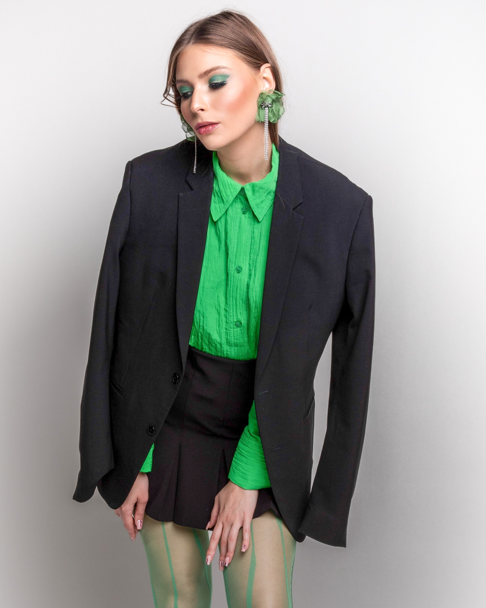 Modelka w czarnej marynarce i zielonej koszuli z błyszczącym, zielonym makijażem oczu i długimi kolczykami z kryształkami.