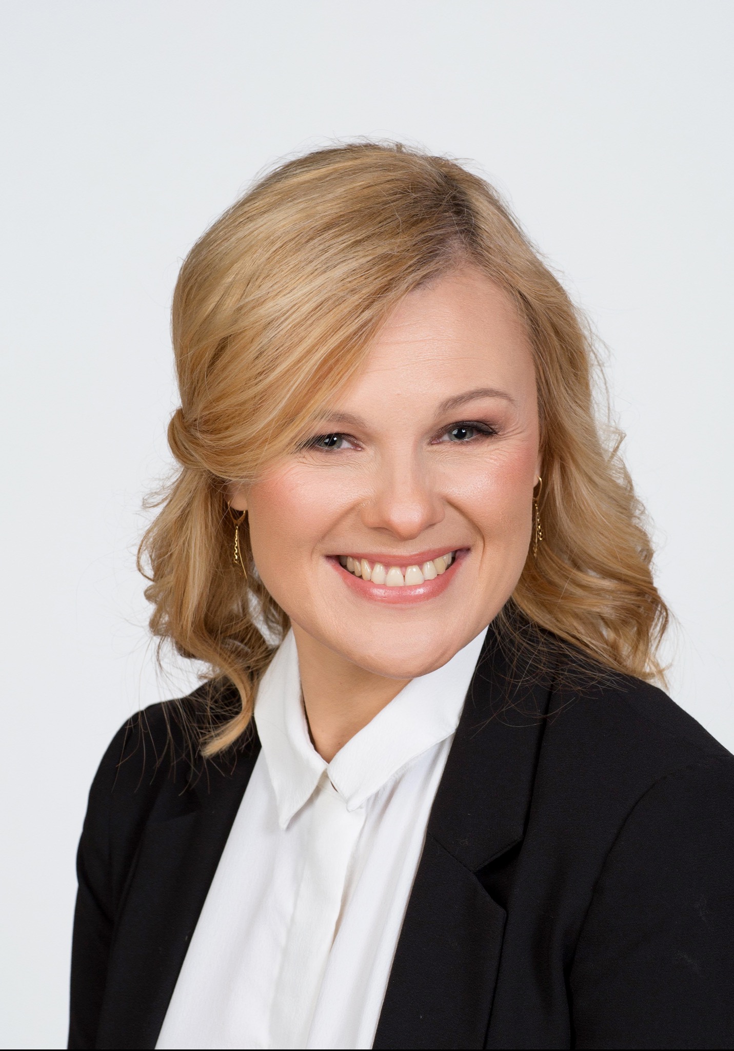 Portret uśmiechniętej blondynki w czarnej marynarce i białej koszuli, z delikatnym makijażem podkreślającym oczy i policzki, oraz upiętymi włosami z lokami opadającymi na ramiona.