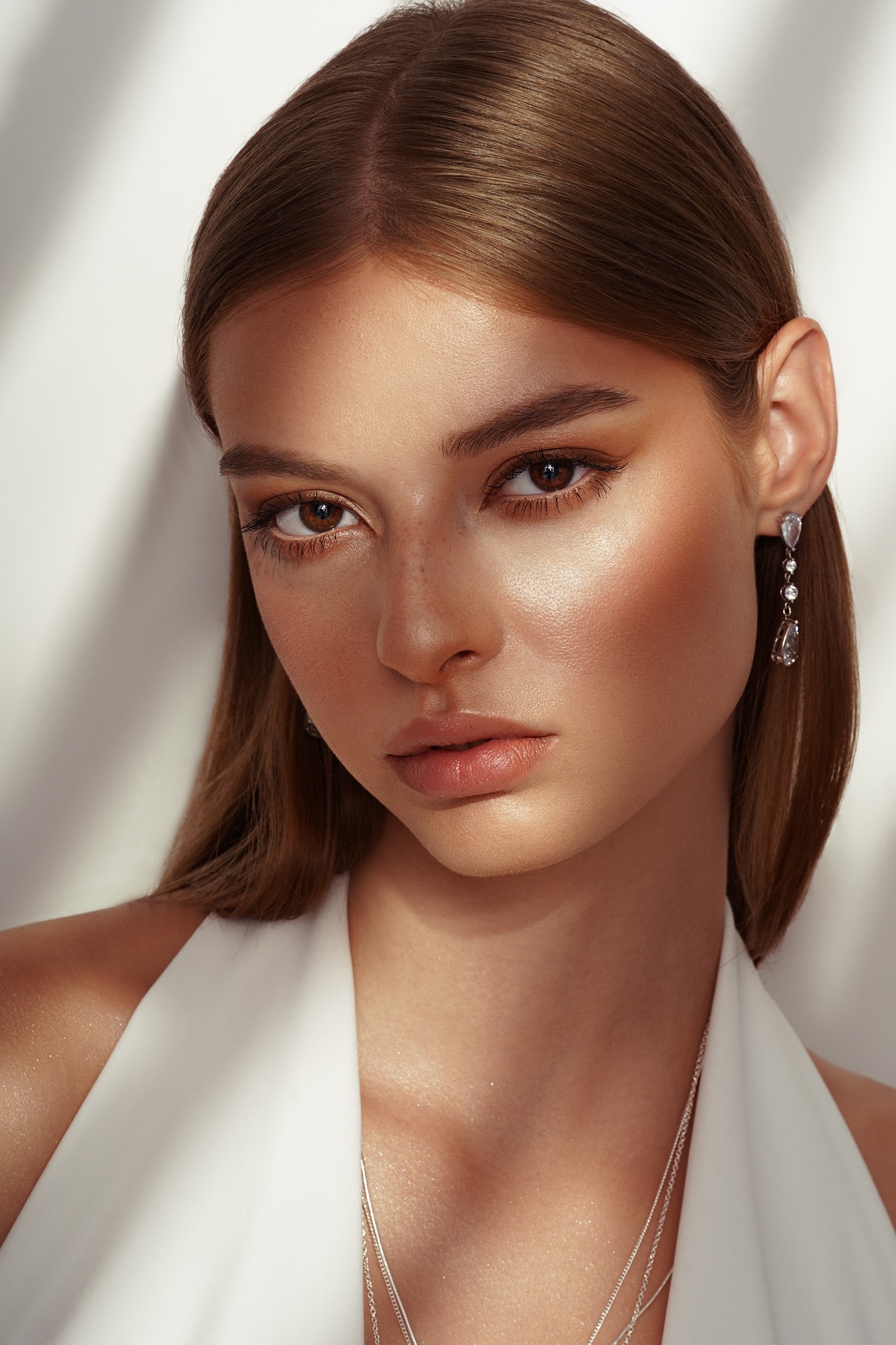 Portret kobiety z subtelnym, brązowym makijażem, podkreślającym kości policzkowe i oczy, z delikatnymi piegami, eleganckimi kolczykami i srebrnym naszyjnikiem, na tle ściany z cieniem.