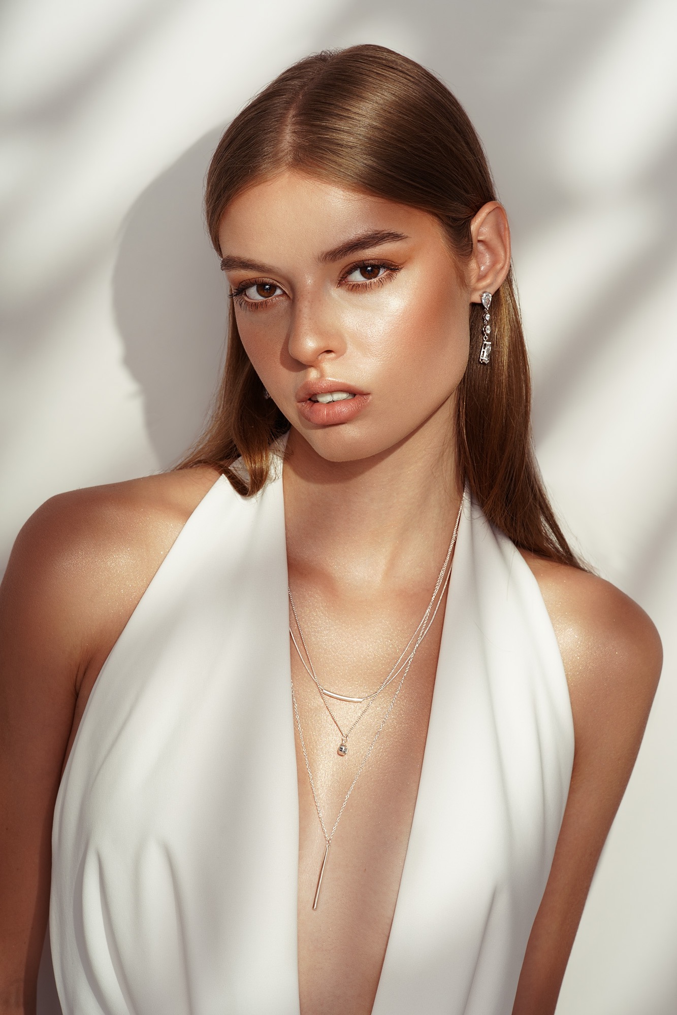 Portret kobiety z delikatnym makijażem, błyszczącą skórą, srebrną biżuterią i białym strojem na tle ściany z cieniem liści.