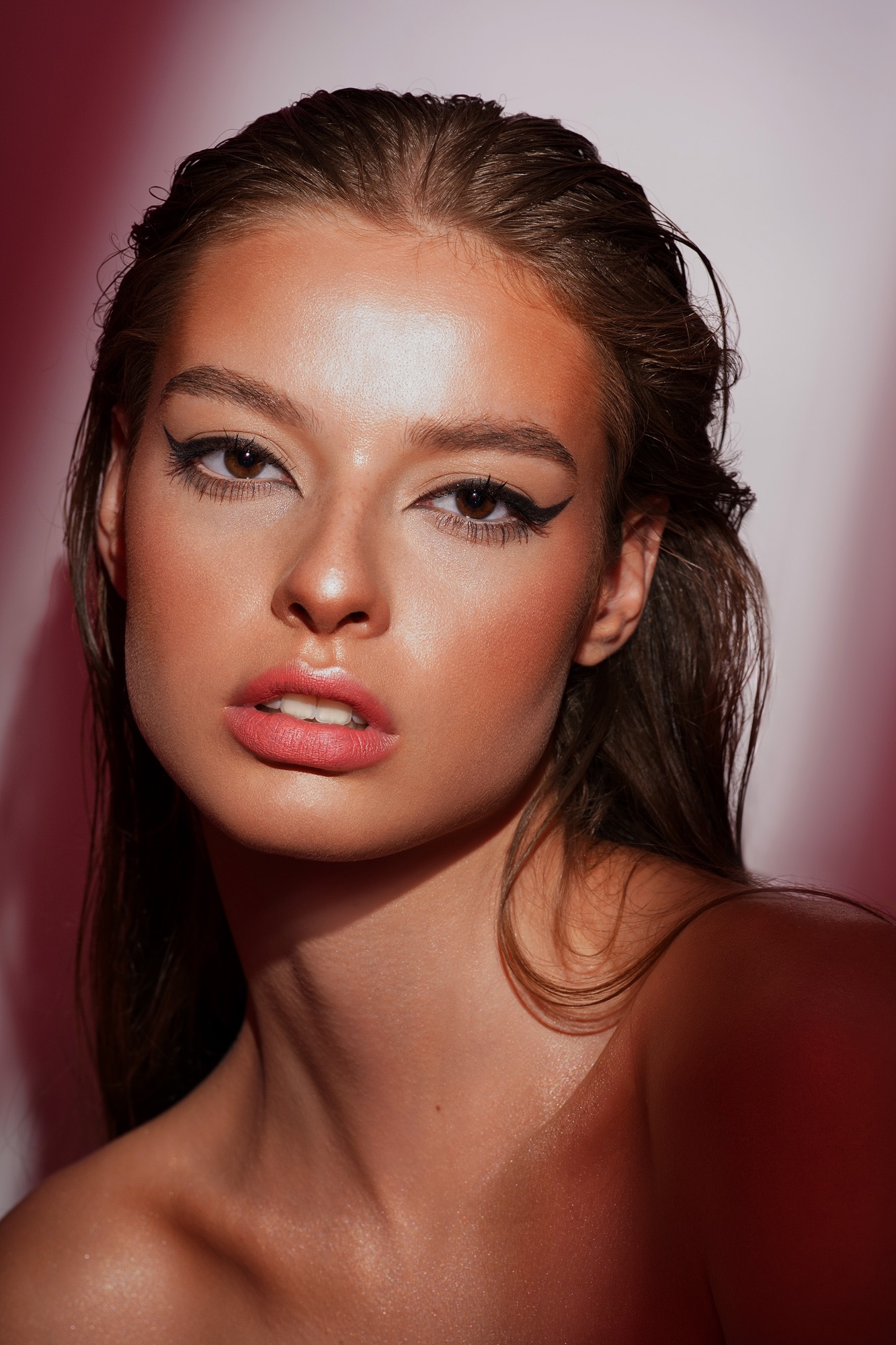 Portret kobiety z profesjonalnym makijażem: wyraźna kreska na powiekach, błyszcząca skóra i mokre włosy, na rozmytym różowym tle.