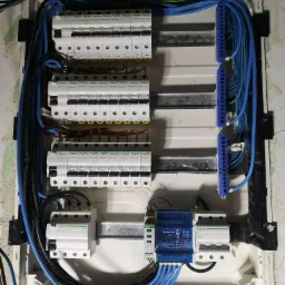 Otwarta skrzynka elektryczna z zainstalowanymi bezpiecznikami Schneider Electric, niebieskie kable ułożone wzdłuż boku, widoczne przewody w różnych kolorach, ściana z widocznymi śladami po malowaniu.